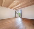 Empty room with wooden flooring and exposed wooden ceiling beams. Features a large window with grey curtains and a sliding glass door leading outside.