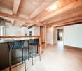 Modern kitchen with exposed wooden beams, bar stools, and a kitchen island. The space features white cabinets, a stainless steel oven, and a sleek countertop.