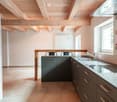Modern kitchen with wooden ceiling beams, stainless steel sink, dark grey cabinets, and a white refrigerator. The space features a bar counter with two chairs and large windows allowing natural light.