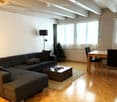 Bright living room with wooden flooring, large windows, grey sectional sofa, coffee table, dining table with chairs, floor lamp, and a small Christmas tree.