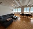 Living room with wooden flooring, exposed wooden ceiling beams, large windows with white curtains, a dark blue sectional sofa, a small coffee table, and a dining area with a wooden table and chairs.