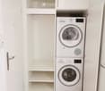A small, modern laundry area with a white washing machine and dryer stacked vertically, white built-in shelving, and wooden flooring.