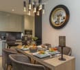 Elegant dining area with a wooden table set for six, surrounded by grey upholstered chairs. The table is adorned with plates of food, wine glasses, and a decorative lamp. A modern kitchen area is visible in the background with sleek cabinets and appliance
