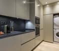 Modern kitchen with built-in oven, microwave, washing machine, and dishwasher. Countertop features a coffee maker, fruits, and a cup.