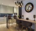Modern kitchen and dining area with sleek cabinets, a central island, bar stools, and a dining table with chairs. Features a large round mirror on the wall and modern lighting.