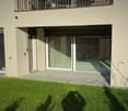 Garden area with a well-maintained lawn, a paved patio, and a modern house entrance with large glass doors and a balcony above.
