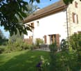 A well-maintained garden area in front of a two-story house with a tiled roof and wooden shutters. The garden features a neatly trimmed lawn, various potted plants, and a small outdoor seating area with a table and chairs.