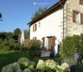 A charming countryside house with a well-maintained garden, featuring a patio area with outdoor furniture, potted plants, and a closed umbrella.
