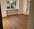 Empty room with wooden flooring, white walls, large windows, radiator, and a small shelf.