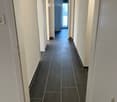 A long, narrow hallway with dark tiled flooring and white walls, featuring several doors and a visible balcony area at the end.