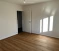 Empty room with wooden flooring, white walls, and large windows allowing natural light. Features a closed door and an open doorway leading to another space.