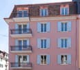 Pink apartment building with multiple balconies and shutters, featuring a tiled roof and classic architectural details.