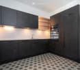 Modern kitchen with dark grey cabinets, white walls, and a hexagonal tile floor. Features include a sink with a modern faucet, built-in lighting, and a built-in oven.