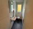 Compact bathroom with a white sink, chrome faucet, toilet, and a small mirror above the sink. The room has light-colored tiles and a dark floor.