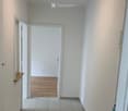 Empty hallway with white walls, tiled floor, wooden floor in the adjacent room, and a ceiling light fixture.