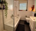 Modern bathroom with a bathtub, toilet, sink, and shower area. Features a black bathroom rug, a plant on a shelf, and a candle on the windowsill.