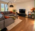 Modern living room with a grey sectional sofa, wooden flooring, a coffee table, a television, and decorative elements including a floor lamp, side table, and shelves.