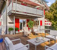 A wooden deck terrace with lounge chairs, a coffee table, and potted plants, featuring a striped awning and a view of the surrounding greenery.