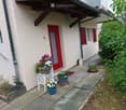 Front entrance of a house with a small garden area, featuring a red front door, a white bench with a flower basket, potted plants, and a small pathway leading to the door.