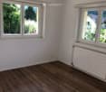 Empty room with wooden flooring, white walls, and large windows offering a view of greenery outside.
