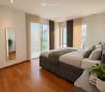 Modern bedroom with wooden flooring, large windows with blinds, a grey upholstered bed with pillows and a green throw, a nightstand with a potted plant, and a mirror on the wall.