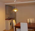 A modern, compact kitchen and dining area with a wooden dining table, white chairs, a washing machine, and a small sink area with a faucet. The space features light-colored cabinets and a hanging pendant light.