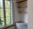 Empty room with white walls, wooden shelves, a window with a view of greenery, and a radiator below the window.