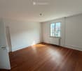 Empty room with wooden flooring, white walls, a window with a view of rooftops, and a radiator below the window.