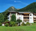 A multi-story white building with balconies and wooden railings, surrounded by green grass and trees, with a mountain view in the background.