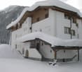 A large, white, multi-story house with a snow-covered roof and wooden accents, situated in a snowy mountain landscape.