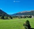 A wide green meadow with scattered trees, a small stream, and a town in the distance surrounded by mountains under a clear blue sky.