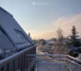 Snow-covered balcony with a bench and chairs, overlooking a snowy residential area with trees and houses.