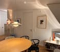 Modern, minimalist room with a round wooden table, black chairs, a small shelf with bottles and a lamp, and a large screen on a stand. The room features light-colored walls, wooden flooring, and several hanging lamps.