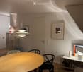 A modern, well-lit room with a round wooden table, black chairs, a small shelf with bottles and a lamp, and a hanging pendant light. The room features white walls, wooden flooring, and a large window allowing natural light to stream in.