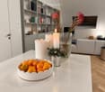 Modern living room with a white marble table, bowl of oranges, candles, vases with flowers, bookshelf, and a grey sofa.