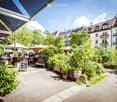 A lush, green outdoor seating area with tables, chairs, and umbrellas, surrounded by potted plants and trees, with multi-story residential buildings in the background.