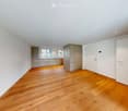 Empty room with wooden flooring, white walls, and a door leading to another room. The adjacent room features a kitchen area with cabinets and a window.