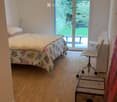 Cozy bedroom with wooden flooring, a bed with floral bedding, a desk with a chair, a lamp, and a suitcase near the window.