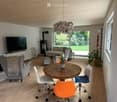 Bright, open-plan living and dining area with light wooden flooring, white walls, and large windows. Features a grey sofa, coffee table, dining table with chairs, and a television on a stand.