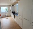 Modern kitchen with white cabinets, wooden flooring, and stainless steel appliances including a stove, sink, and fridge. A small table with a chair is placed near the window.