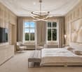 Elegant bedroom with a large bed, bench at the foot, two armchairs, dresser, television, and artwork on the wall. The room features large windows with curtains and a modern chandelier.
