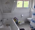 Bathroom with a bathtub, toilet, and a small child's stool. The walls are tiled with fish decals, and there is a window with blinds. A shower curtain with a sea theme is visible.