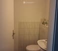 Small bathroom with a toilet, sink, and a small shelf holding a cup, soap dispenser, and toilet paper. The walls are tiled with beige tiles, and the floor is also tiled.