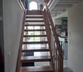 Wooden staircase with a wooden handrail, leading to a bright room with a large arched window and visible kitchen area in the background.