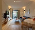 Bright living room with wooden flooring, a sofa, coffee table, dining table, and a TV. Large sliding doors open to a balcony.
