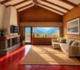 Cozy living room with exposed wooden beams, large window offering mountain view, stone fireplace, cream sectional sofa, coffee table, and potted plants on windowsill.