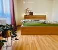 Cozy bedroom with wooden flooring, a bed with green bedding, a nightstand with picture frames, and a potted plant near the window.