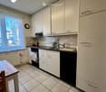 Modern kitchen with white cabinets, stainless steel sink, dishwasher, and a small dining area with a table and chair. The space features tiled flooring and a window with a view of trees.