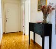 Hallway with wooden flooring, a small black table with a vase of dried flowers and a decorative plate, and a framed picture on the wall.