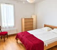 A well-lit bedroom with a wooden bed, red blanket, wooden dresser, and a red chair near the window.